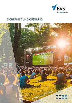 Auf einer Wieder mit hohen Bäumen ist eine große beleuchtete Leinwand aufgespannt, die ein Fußballspiel zeigt. Auf dem Rasen davor sieht man viele Menschen in Sommerkleidung sitzen.