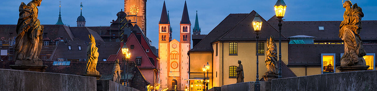 Der Bildausschnitt zeigt den abendlichen Blick von der alten Mainbrücke auf den Dom. Die Laternen an der Brück entlang leuchten schon.