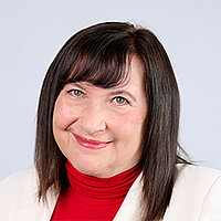 Michaela Wintermayr-Greck Eine Frau mit längeren dunklen Haaren hat blaue Augen und trägt einen weißen Blazer über einem roten Pullover.