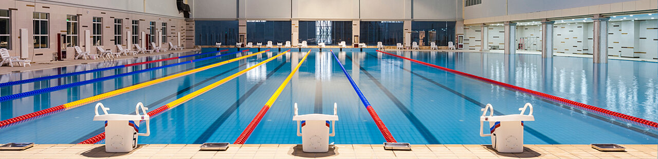 Im Vordergrund sieht man 3 Springblöcke dahinter ein langes Schwimmbecken in einer großen Halle mit abgetrennten Schwimmbahnen.