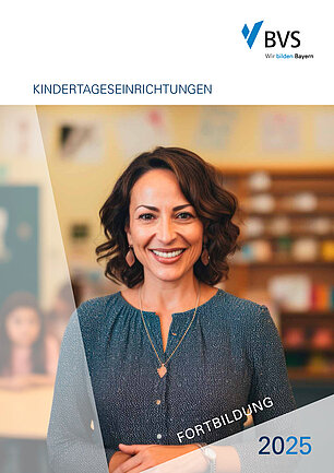 Vor einem unscharfen Hintergrund mit Regal und Kindern steht eine dunkelhaarige Frau mittleren Alters. Sie lächelt und trägt ein dunkelblau gemustertes Oberteil mit einem runden Ausschnitt. 