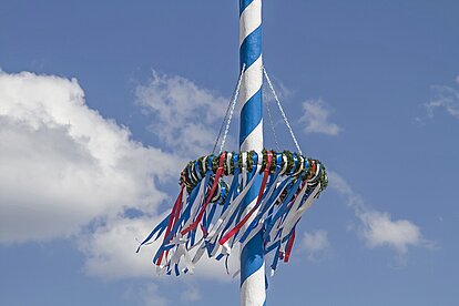 Es ist ein Stück eines bayerischen Maibaums zu sehen, an dem ein Blumenkranz hängt.