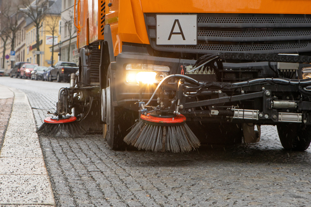 Aus der Untersicht sieht man vom Straßenrand aus den linken unteren Teil einer Kehrmaschine mit beleuchteten Scheinwerfer.